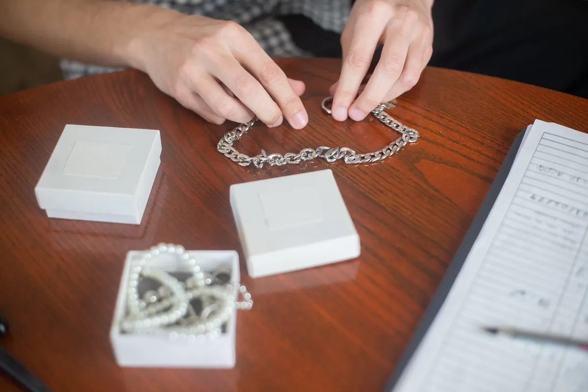 Hands arranging a silver chain necklace on a wooden table with jewelry boxes, reflecting the selling process.